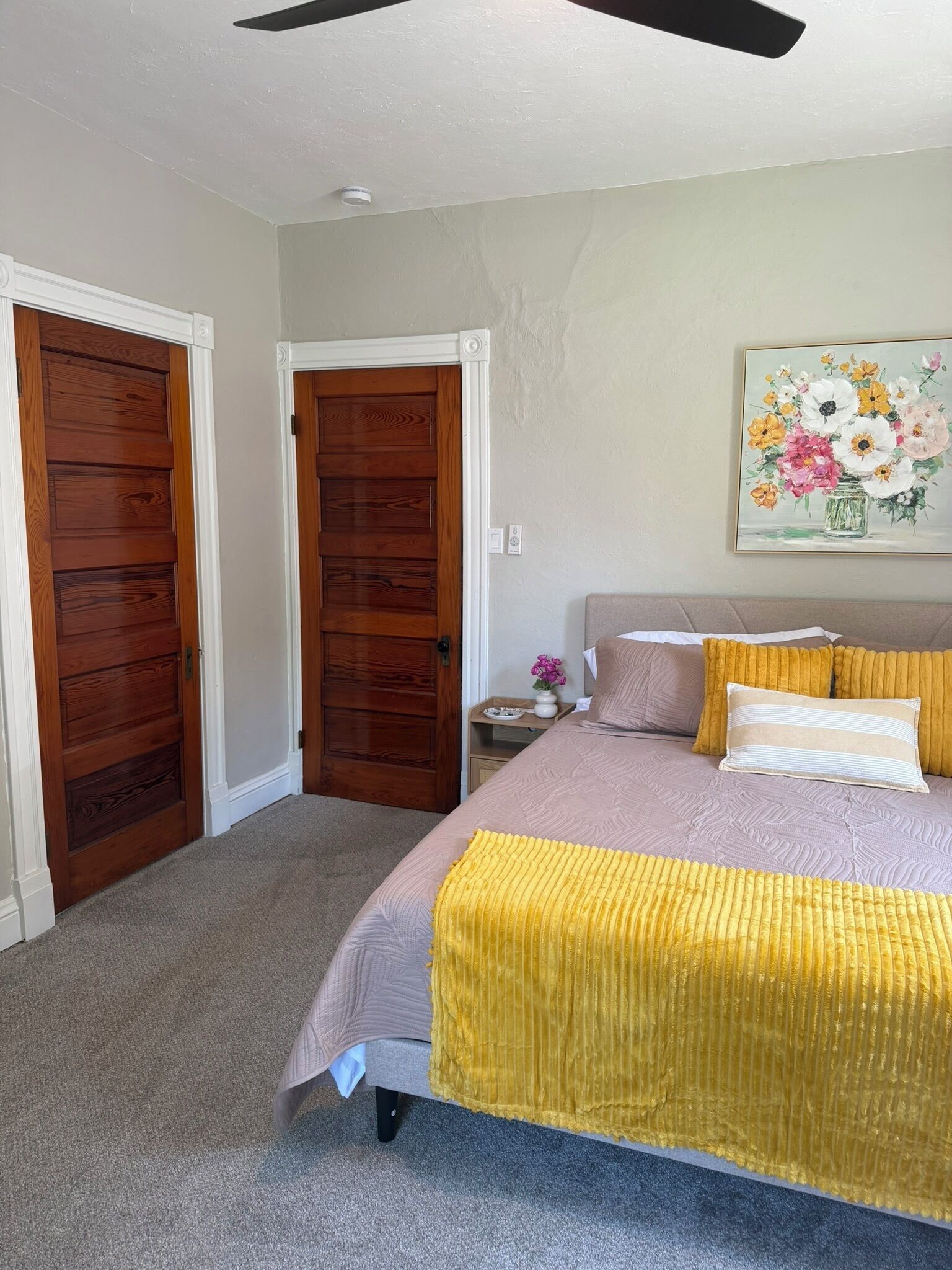 Une chambre à coucher avec un lit, deux portes en bois, une table de chevet, un tableau et un ventilateur de plafond.