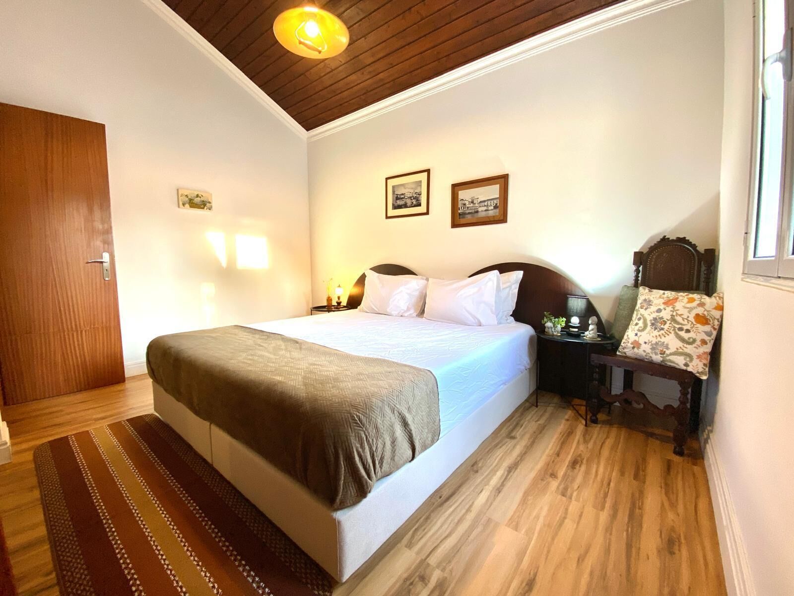Habitación de hotel con cama, suelo de madera, mesita de noche, una silla y una ventana.