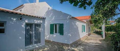 A white building with a red-tiled roof, green shutters, and a narrow pathway leading to a door.