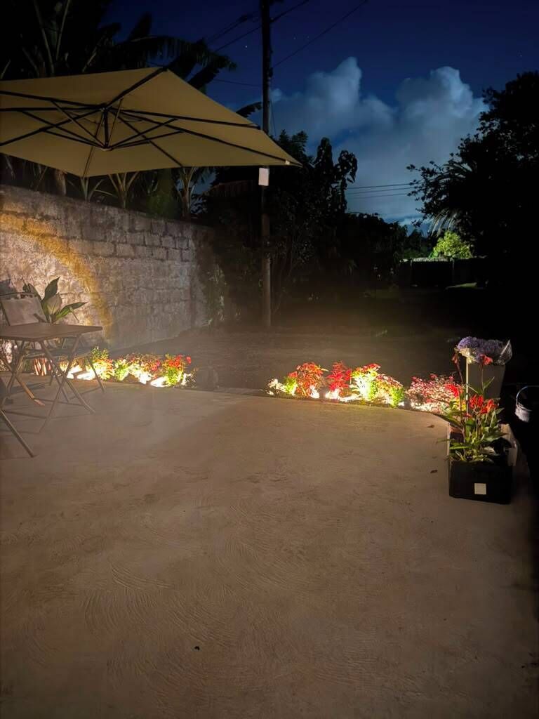 An outdoor area with a concrete floor, a stone wall, and a large umbrella. There are potted plants and flowers along the wall.
