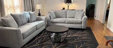 A living room with two grey sofas, a round coffee table, a black rug with a leaf pattern, a lamp, and a mirror on the wall.