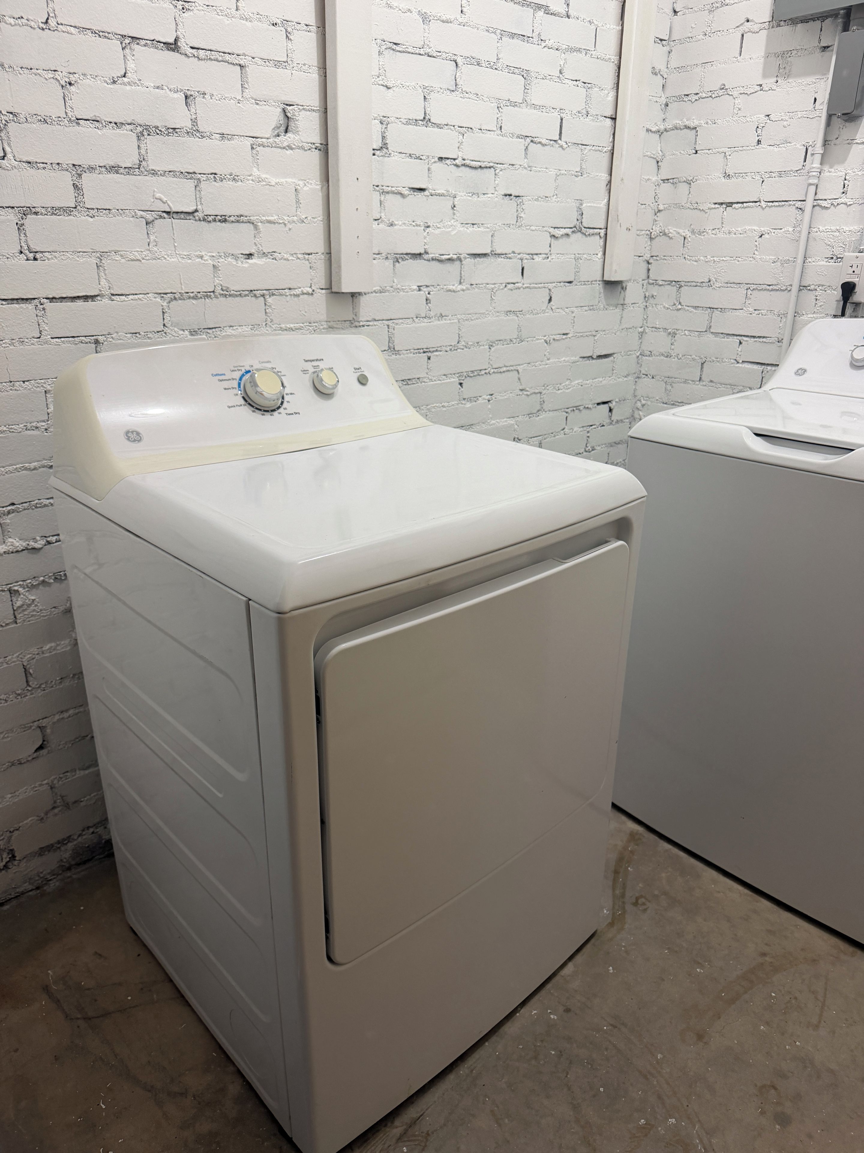 A front-loading washing machine in a laundry room with a white brick wall.