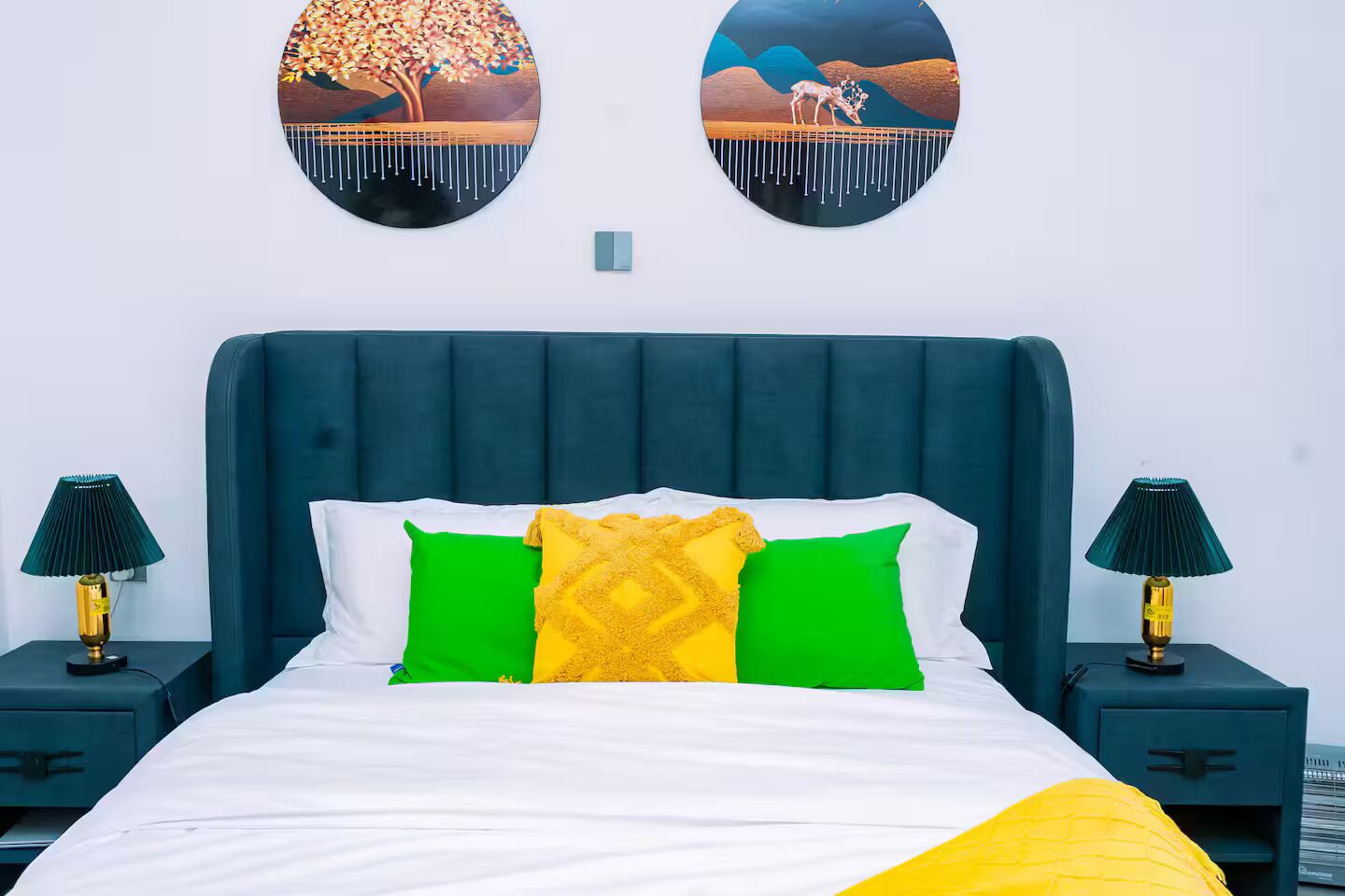 A bed with a dark headboard, white bedding, and green and yellow pillows. There are two bedside tables with lamps and a wall-mounted light switch.