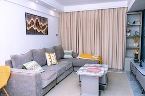 Ein modernes Wohnzimmer mit einem grauen Ecksofa, einem Marmor-Couchtisch und einem großen Fenster mit beigen Vorhängen.