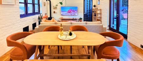 A modern dining area with a wooden table and orange chairs, a television in the background, and a view of the outdoors through large windows.