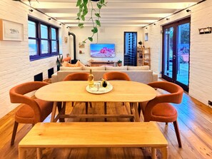 A modern dining area with a wooden table and orange chairs, a television in the background, and a view of the outdoors through large windows. - Curated sanctuary in the heart of the city (Cape Town)