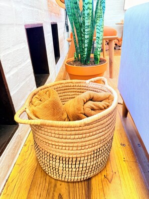 A wicker basket with towels on a wooden floor, a potted plant, and a white wall with a window. - Curated sanctuary in the heart of the city (Cape Town)