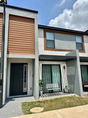 A modern two-story residential building with a flat roof, large windows, and a covered outdoor seating area. - Magical Family Escape! Spacious 3BR Home Minutes from Disney! (Kissimmee)