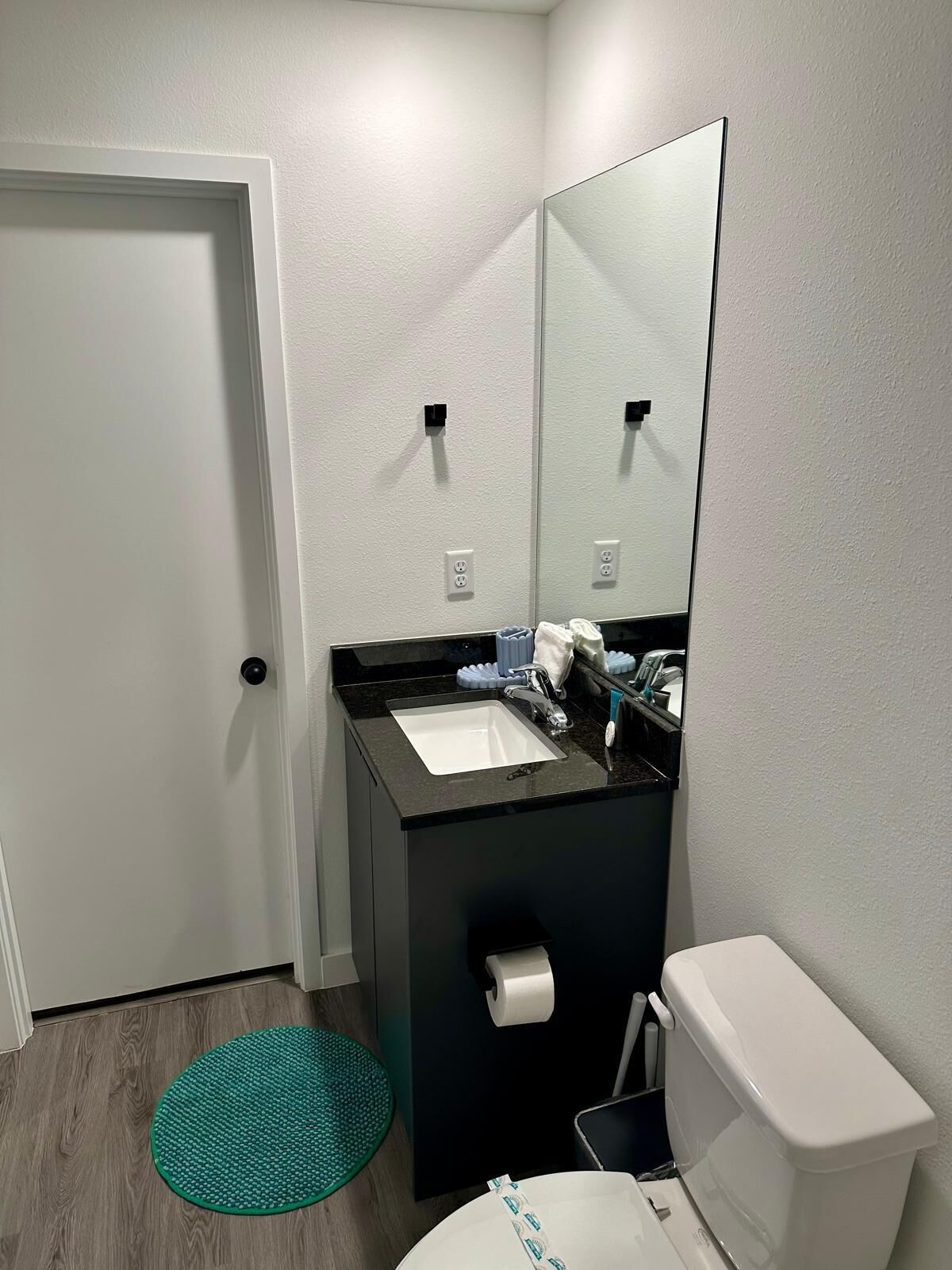 Un baño con inodoro blanco, lavamanos negro, espejo y un tapete verde en el suelo.