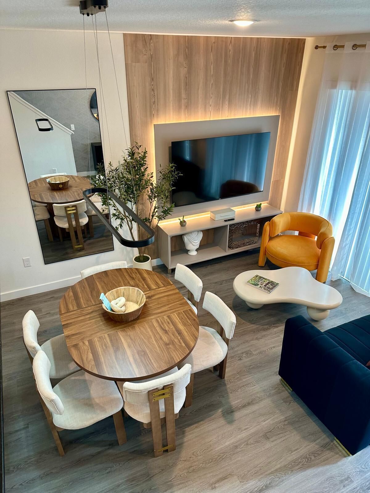 A modern living room with a round wooden dining table, white upholstered chairs, a flat-screen TV, a sofa, and a potted plant.