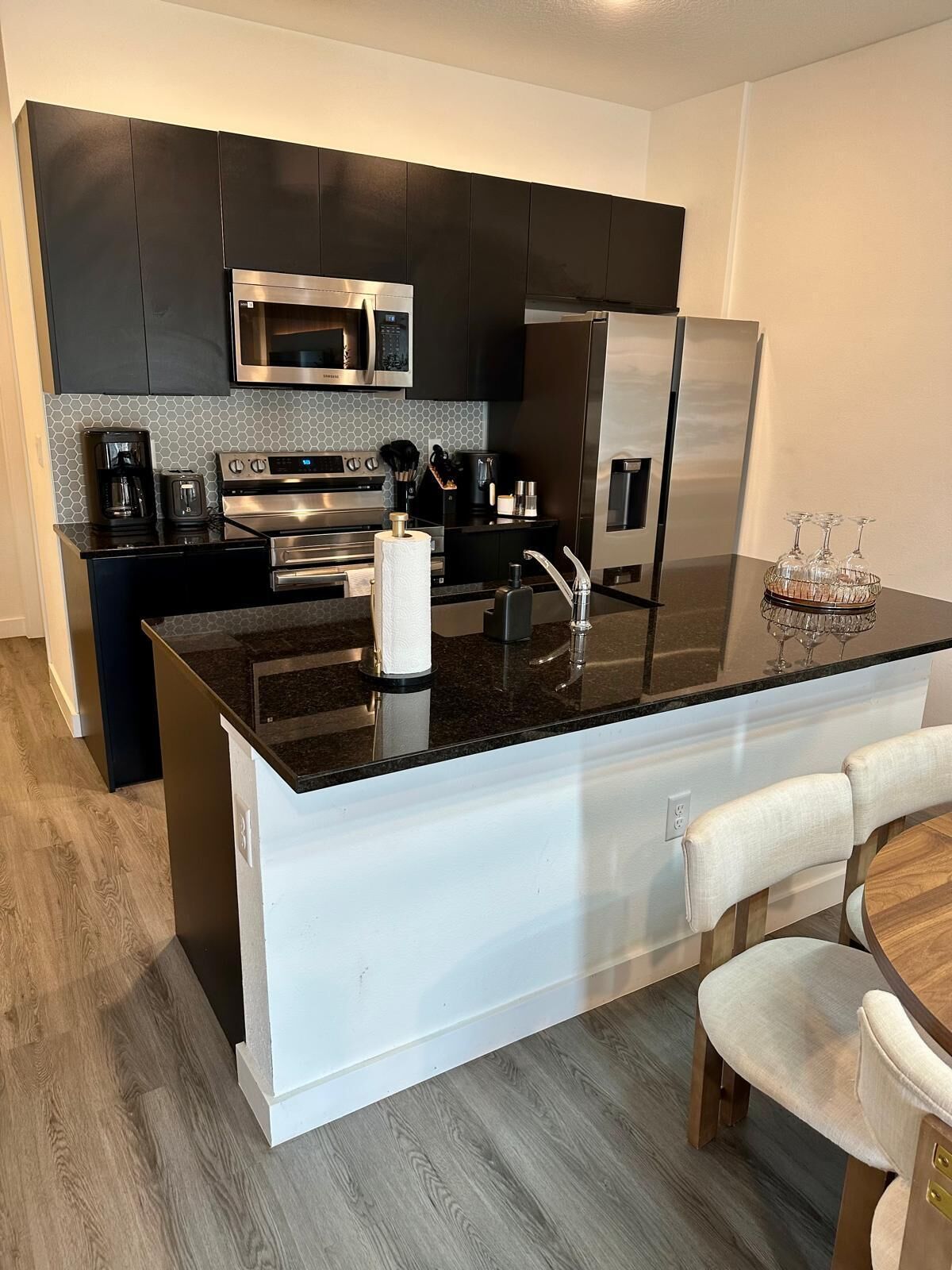 Una cocina moderna con armarios oscuros, electrodomésticos de acero inoxidable, una isla central con encimera negra y un comedor con mesa de madera y sillas blancas.