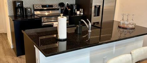 Una cocina moderna con armarios oscuros, electrodomésticos de acero inoxidable, una isla central con encimera negra y un comedor con mesa de madera y sillas blancas.