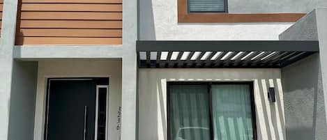 Un edificio residencial moderno de dos pisos con una puerta principal, un pequeño porche con un banco y una planta en maceta.