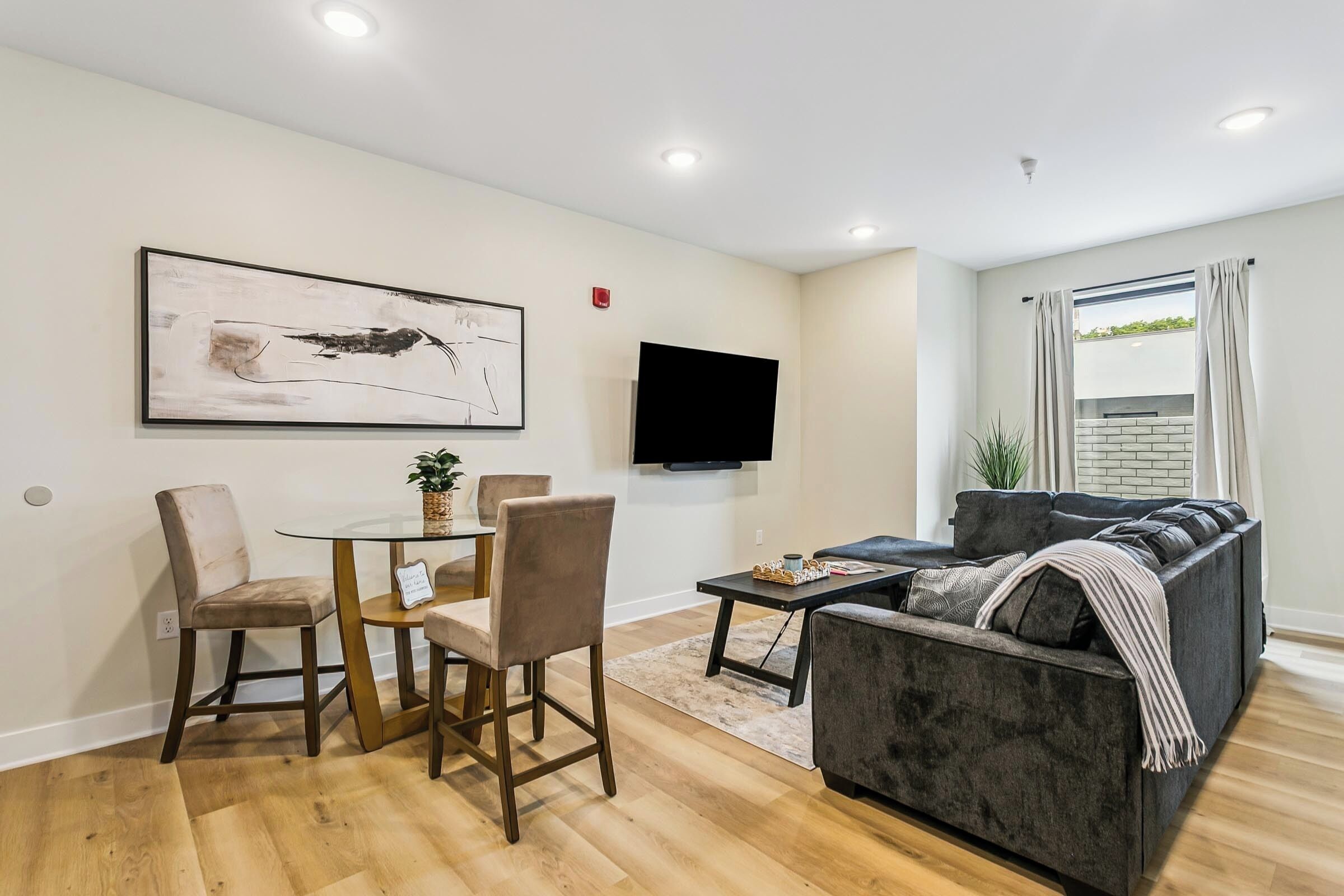 A modern living room with a sofa, a dining table with chairs, a flat-screen TV mounted on the wall, and a large abstract painting.
