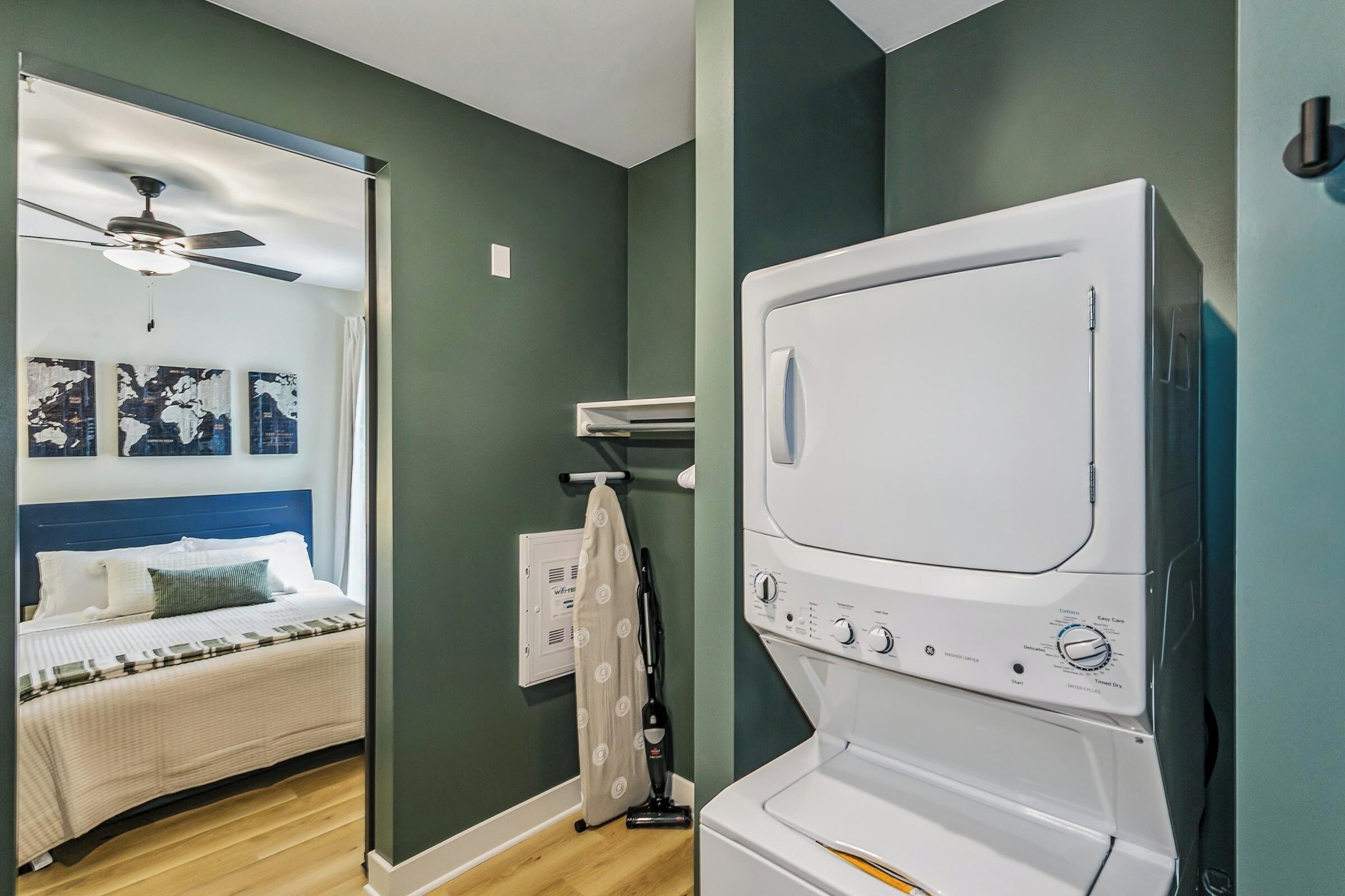 A hotel room with a bed, a ceiling fan, a washing machine, and a view of the outside.