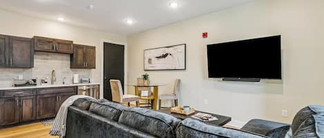 Ein modernes Wohnzimmer mit einem grauen Ecksofa, einem flachbildfernseher an der Wand und einer Küchenzeile mit dunklen Holzschränken und einer weißen Arbeitsplatte.