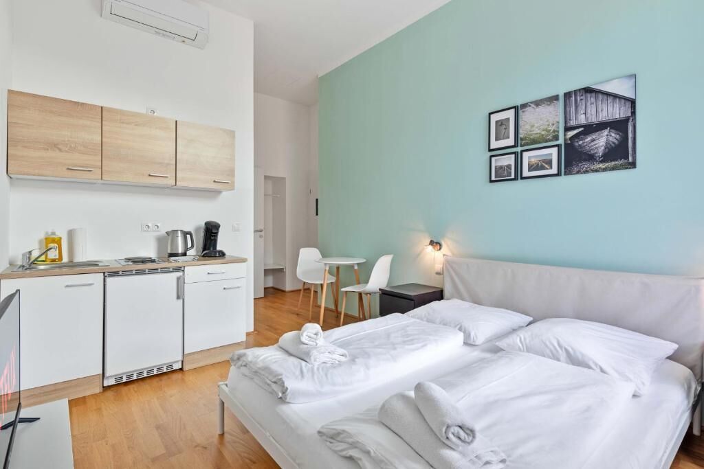 A hotel room with a bed, a small table, a chair, a kitchenette, and a wall with pictures.