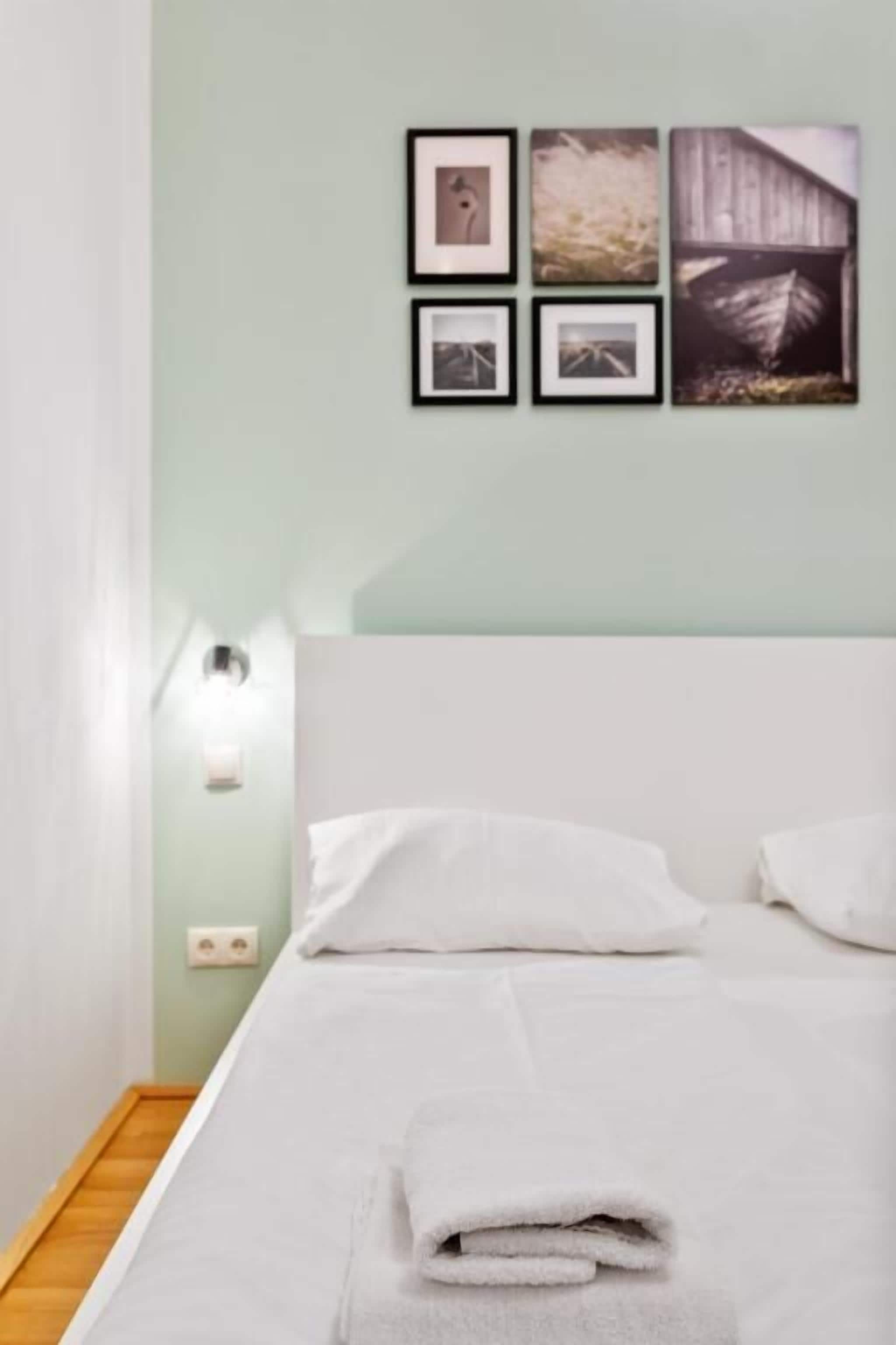 Un letto rifatto con cura, un asciugamano piegato, una parete di colore chiaro e una serie di quadri appesi sopra.
