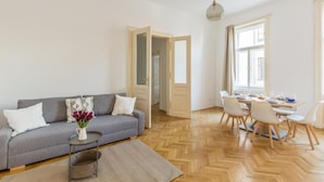 A modern living room with a grey sofa, a small side table with a vase, a dining area with a table set for four, and a herringbone wooden floor. - Cityswap- Wien Naschmarkt (Wien)