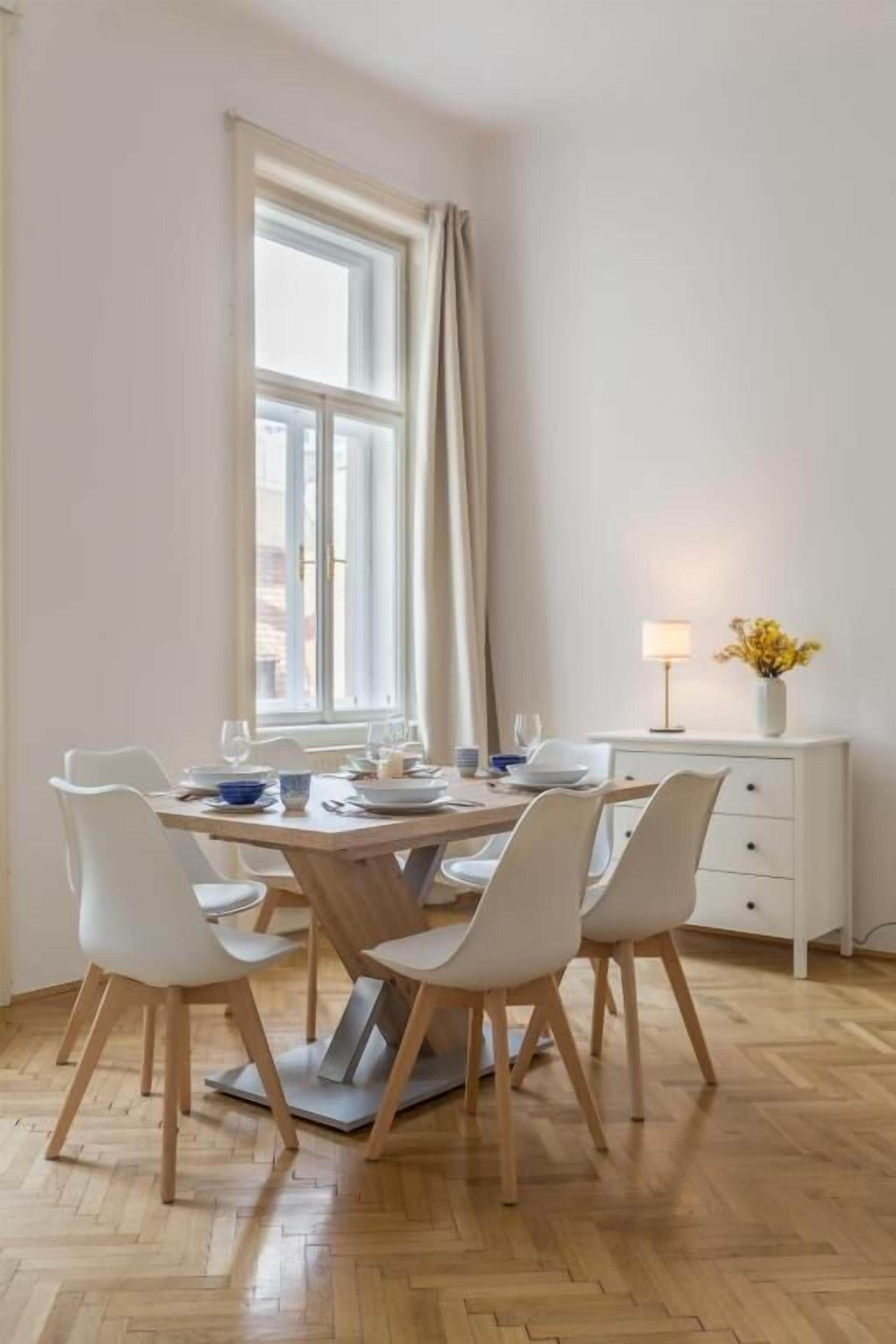 Un comedor con una mesa redonda, sillas blancas, una ventana con cortinas, una lámpara y un jarrón con flores.