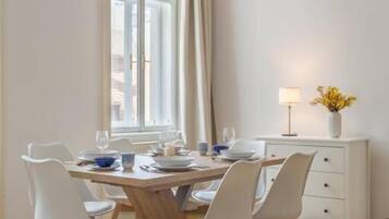 Un comedor con una mesa redonda, sillas blancas, una ventana con cortinas, una lámpara y un jarrón con flores.