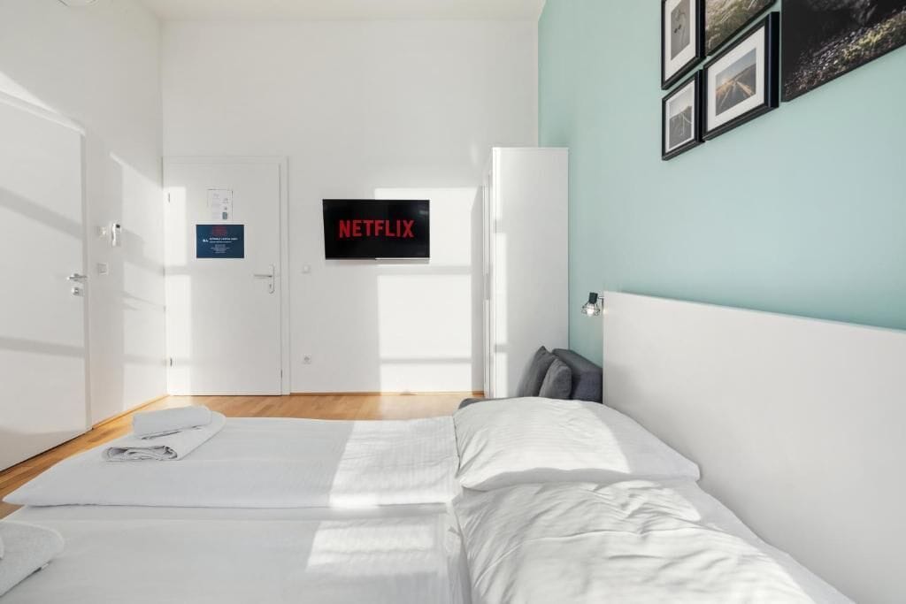Hotellihuone, jossa on sänky, ovi, jossa on kyltti, seinälle asennettu televisio ja kehystetyt valokuvat.