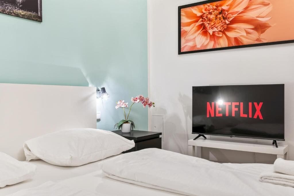 Una camera d'albergo con un letto, un comodino con un vaso di fiori, un televisore che mostra 'NETFLIX' e un quadro floreale appeso al muro.