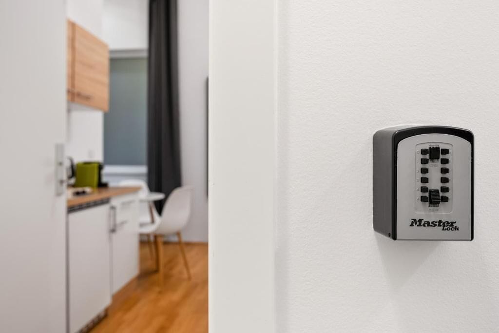 A Master Lock device on a white wall in a room with a wooden floor, white furniture, and a kitchen area in the background.