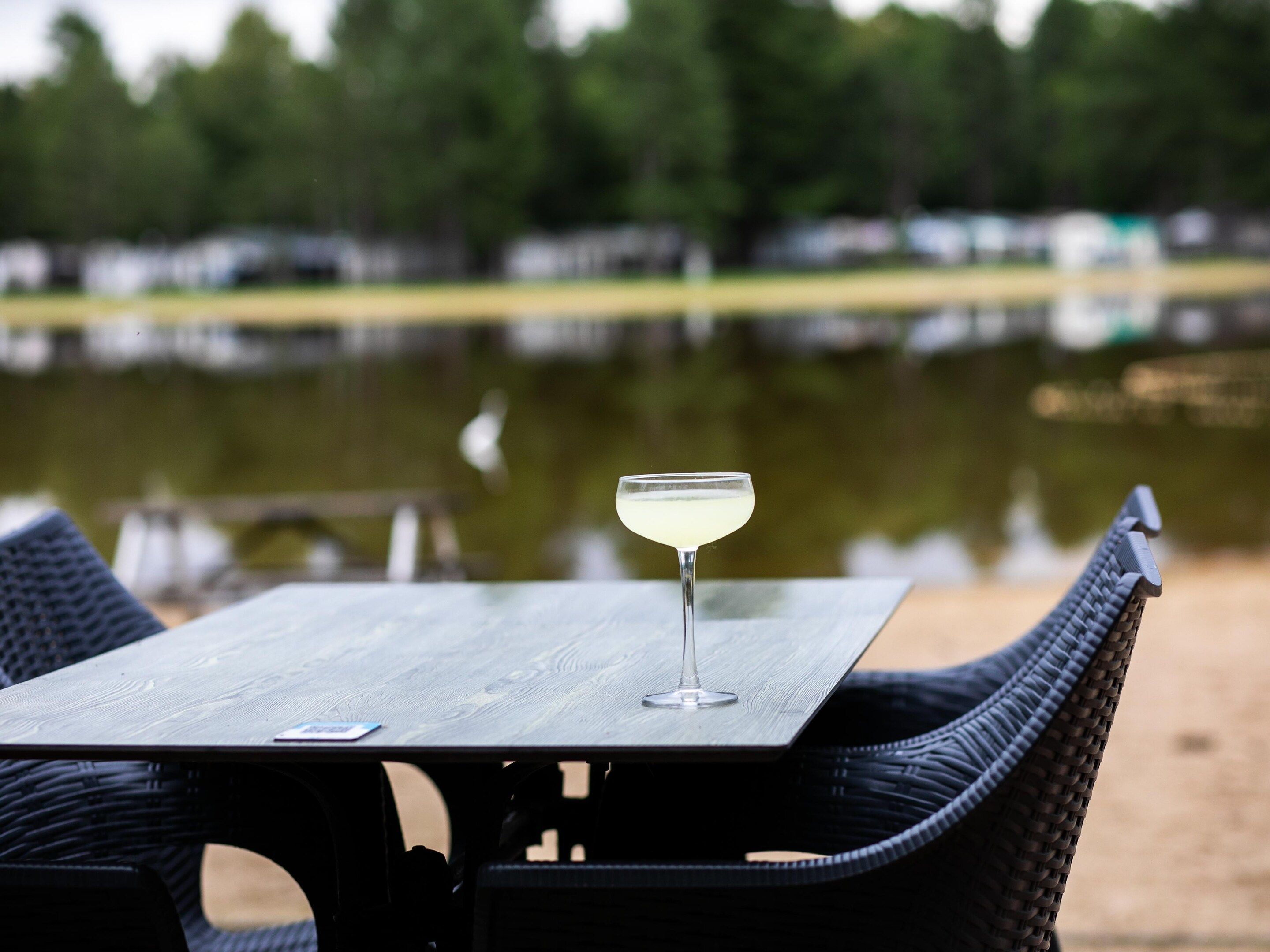 Une table avec un verre et des chaises, placée devant un paysage composé d’une étendue d’eau et d’arbres.