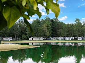 Un lago tranquilo con un solo ave blanca, rodeado de árboles densos y una hilera de viviendas móviles.