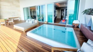 Ein moderner Poolbereich im Freien mit Holzterrasse, Liegestuhl und einem Wohnraum mit Glasfassade.