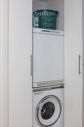 A compact laundry room with a stacked washer and dryer, a green laundry basket on top, and a white wall. - The Dandy Relaxed Living near St Kilda Beach and CBD (Caulfield North)