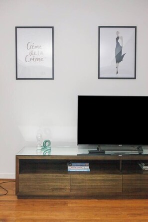 A room with a flat-screen TV on a wooden TV stand, two framed artworks on the wall, and a glass coffee table with a green object on it. - The Dandy Relaxed Living near St Kilda Beach and CBD (Caulfield North)