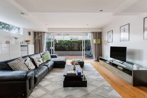 Ein modernes Wohnzimmer mit einer schwarzen Ledersofa, einem Couchtisch, einem Flachbildfernseher und einer Glasschiebetür, die auf einen mit Grünpflanzen bepflanzten Balkon führt.