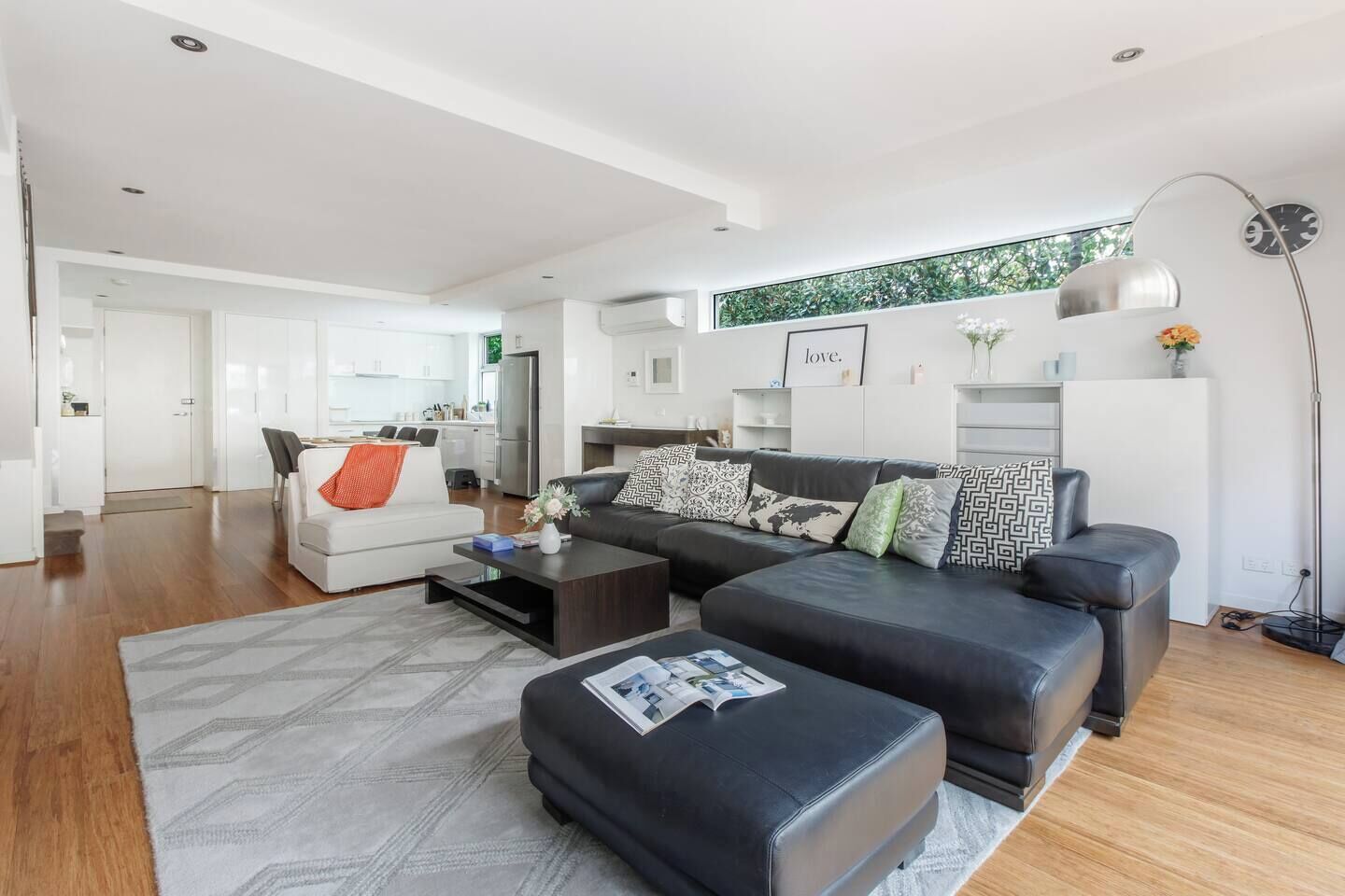 A modern living room with a sofa, coffee table, and ottoman, a kitchen in the background, and a large window with a view of greenery.