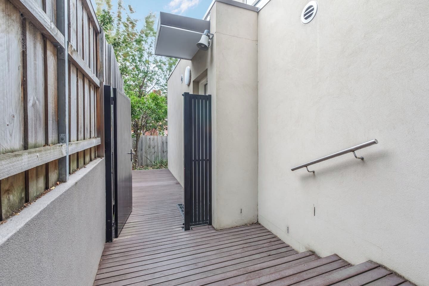 A narrow outdoor corridor with a wooden deck, a black gate, and a white wall.