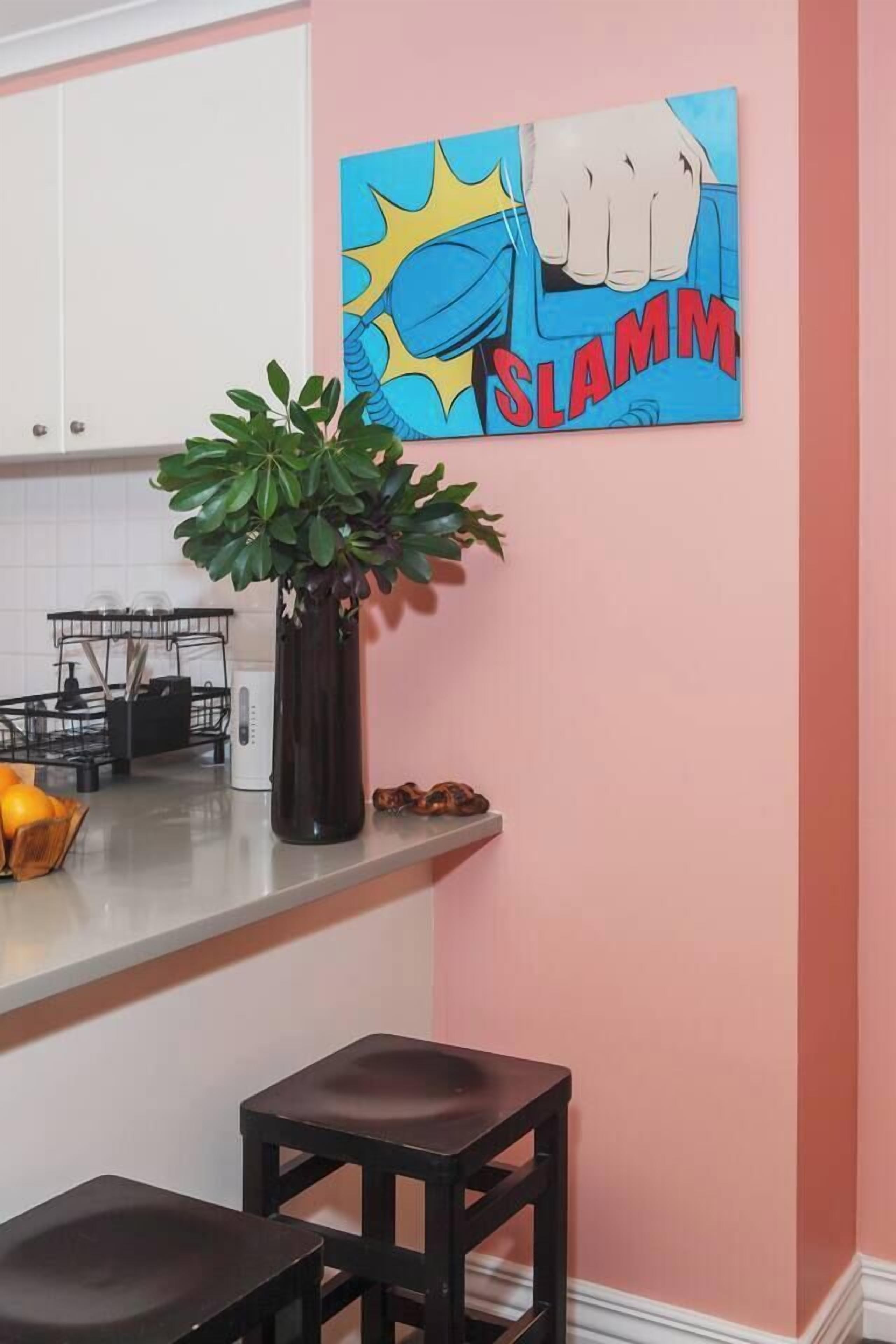 A kitchen with a pink wall, a black stool, a plant in a black vase, and a comic book-style painting.