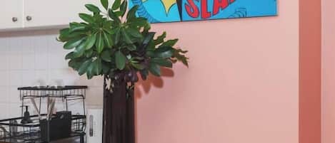 A kitchen with a pink wall, a black stool, a plant in a black vase, and a comic book-style painting.