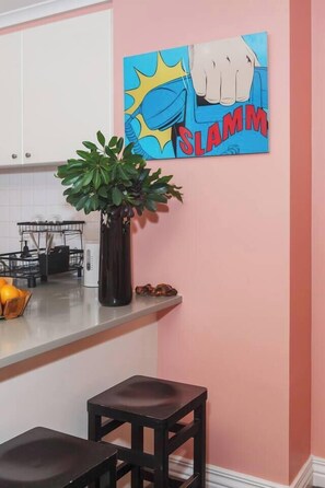 A kitchen with a pink wall, a black stool, a plant in a black vase, and a comic book-style painting. - Coral Atelier Bright Artistic Hideaway in the CBD (Melbourne)