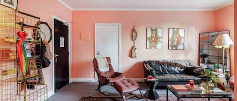 A living room with a sofa, armchair, and a coffee table. There is a door, a mirror, and a wall with artwork.
