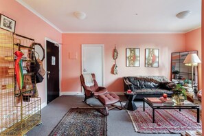 Ein Wohnzimmer mit Sofa, Sessel und Couchtisch. Eine Tür, ein Spiegel und eine Wand mit Kunstwerken sind zu sehen.