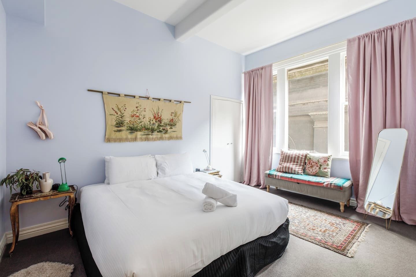 Un dormitorio con una cama grande, una mesita de noche, una lámpara de noche, una ventana con cortinas, un banco con cojines, un espejo y un tapiz en la pared.