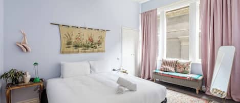Un dormitorio con una cama grande, una mesita de noche, una lámpara de noche, una ventana con cortinas, un banco con cojines, un espejo y un tapiz en la pared.