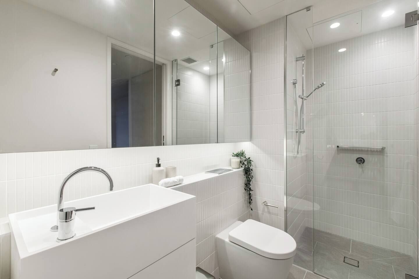 A modern bathroom with a large mirror, a white sink, a toilet, and a glass-enclosed shower.