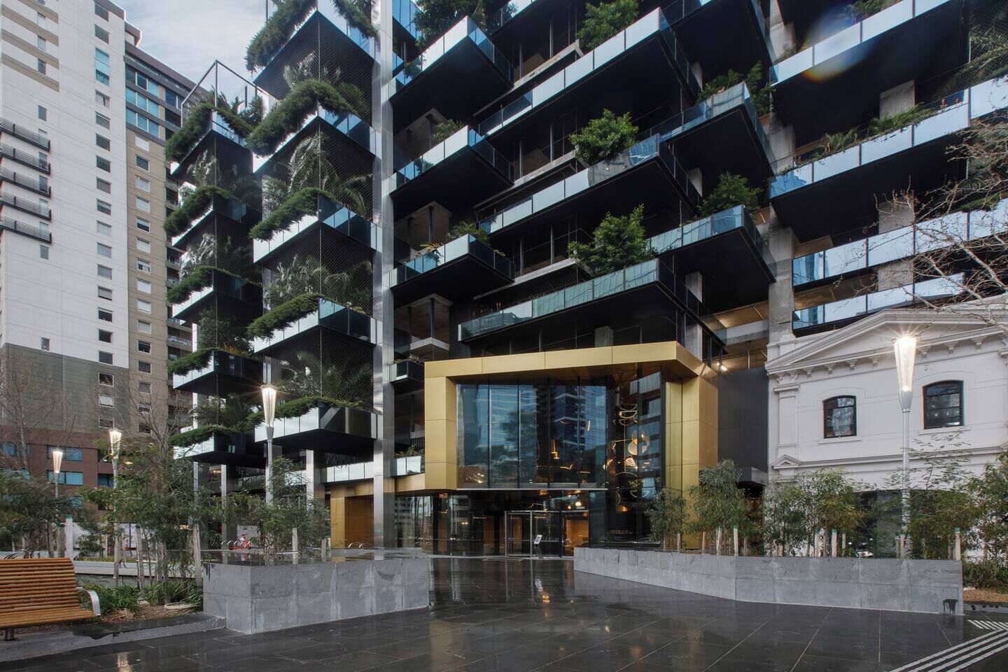 A modern multi-story building with balconies, a central entrance, and surrounding greenery.