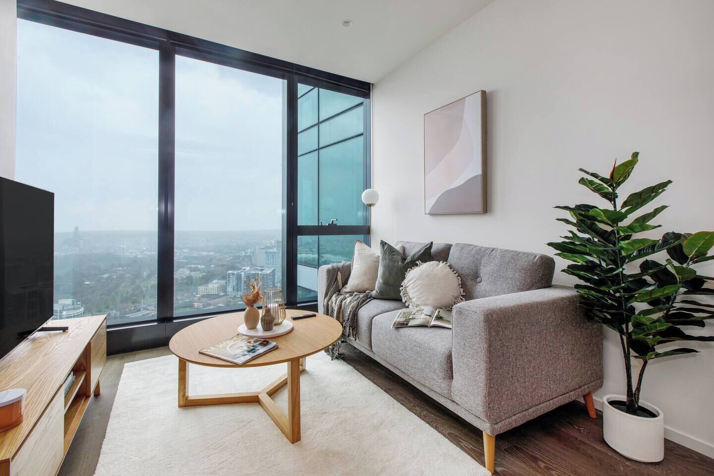 Ein modernes Wohnzimmer mit großem Fenster, einem grauen Sofa, einem runden Couchtisch und einer Topfpflanze.