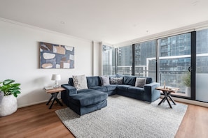A modern living room with a sectional sofa, a large window, and a city view. - Sentinel Vista Urban Stay with Pool and Tennis Court (Southbank)