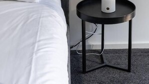 En säng med vita sängkläder och en grå sänggavel, ett runt svart sidobord med en vit lampa och ett grått mattbeklätt golv.