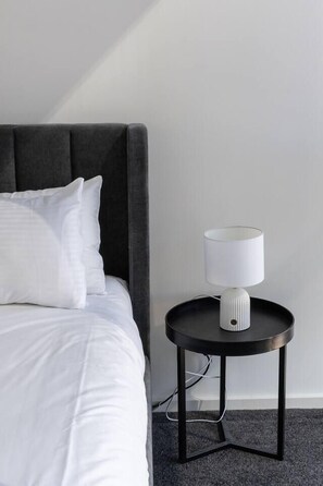 A bed with white bedding and a grey headboard, a round black side table with a white lamp, and a grey carpeted floor. - Sentinel Vista Urban Stay with Pool and Tennis Court (Southbank)