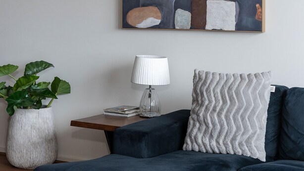 Ett modernt vardagsrum med en mörkblå hörnsoffa, en vit, strukturerad prydnadskudde, ett litet sidobord med en lampa och en krukväxt.